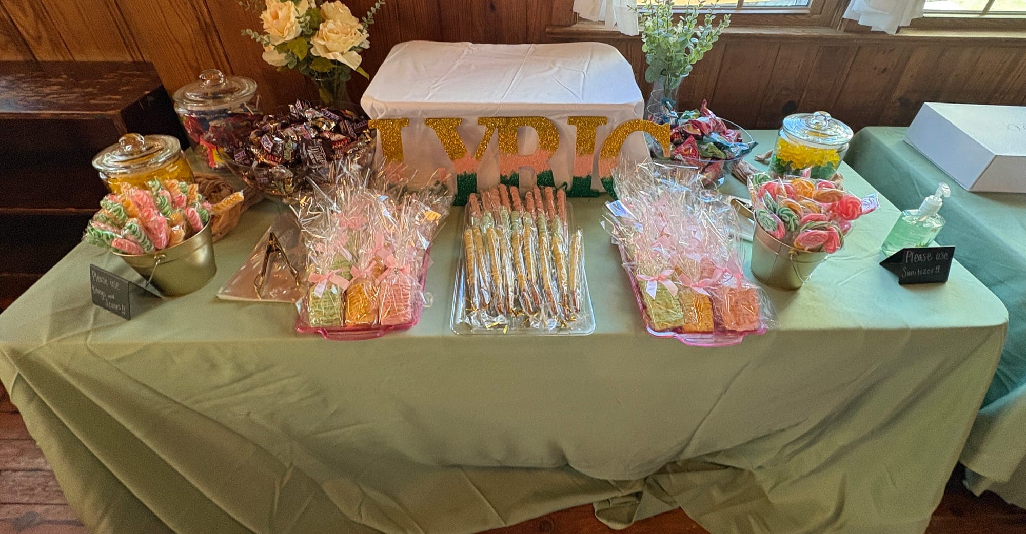 Styled candy buffet table with apothecary jars of candy, swirl lollipops, wrapped rice crispy treats and chocolate-covered pretzel rods on a sage green tablecloth